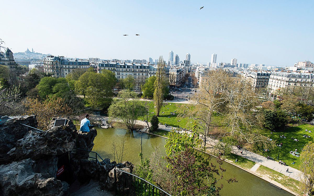 Aerial view of Parc des Buttes-Chaumont in Paris with city skyline visible in the distance.