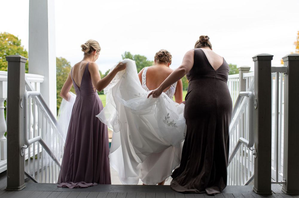 Three figures in elegant dresses stand on a white porch, lifting the bride's gown.