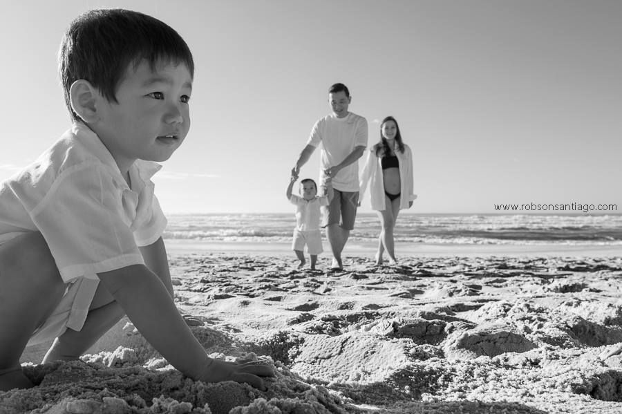A black and white beach scene with a child playing in the sand while two people walk in the distance near the ocean.