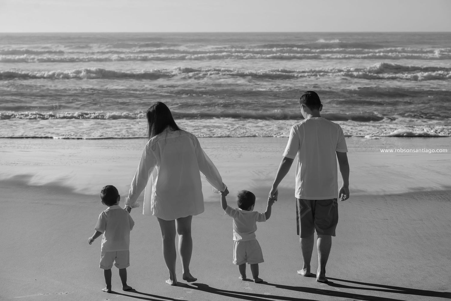 A family silhouette holds hands while walking along the shoreline of a sandy beach during sunset.