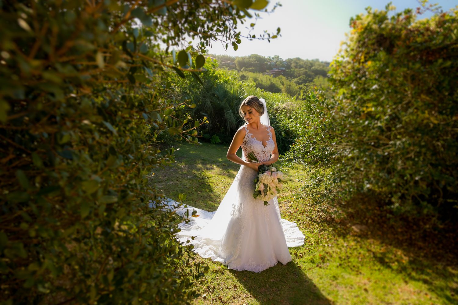 A bride in a white wedding gown holding a bouquet stands in dappled sunlight between garden hedges.