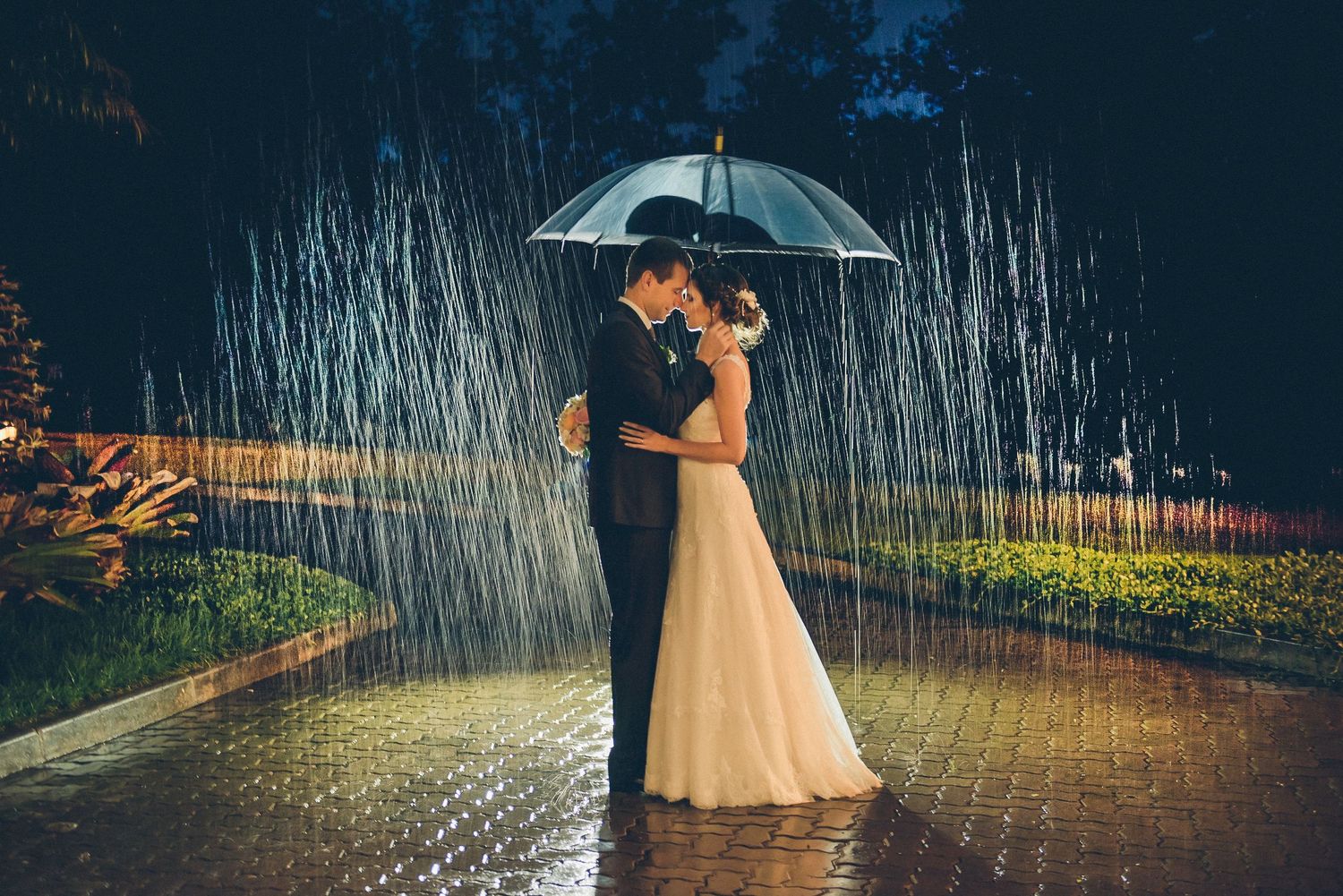 A couple shares an intimate moment under an umbrella in the rain with evening lights creating a magical glow.