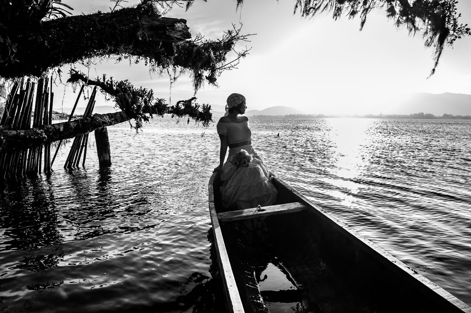 A black and white photo of someone paddling a canoe at sunset with overhanging trees framing the scene.