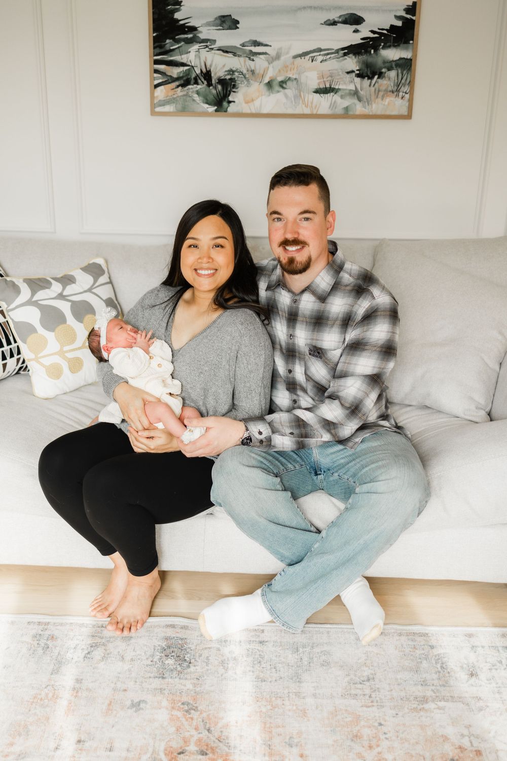 A couple sits on a gray couch with their newborn baby beneath landscape artwork.