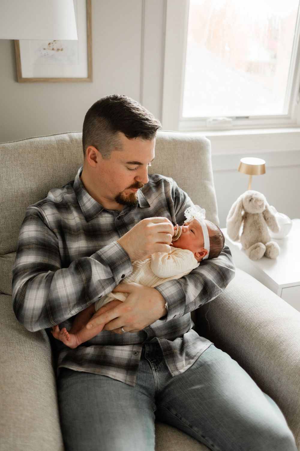 A tender moment in a nursery as a caregiver in plaid feeds a tiny infant in a cozy armchair.