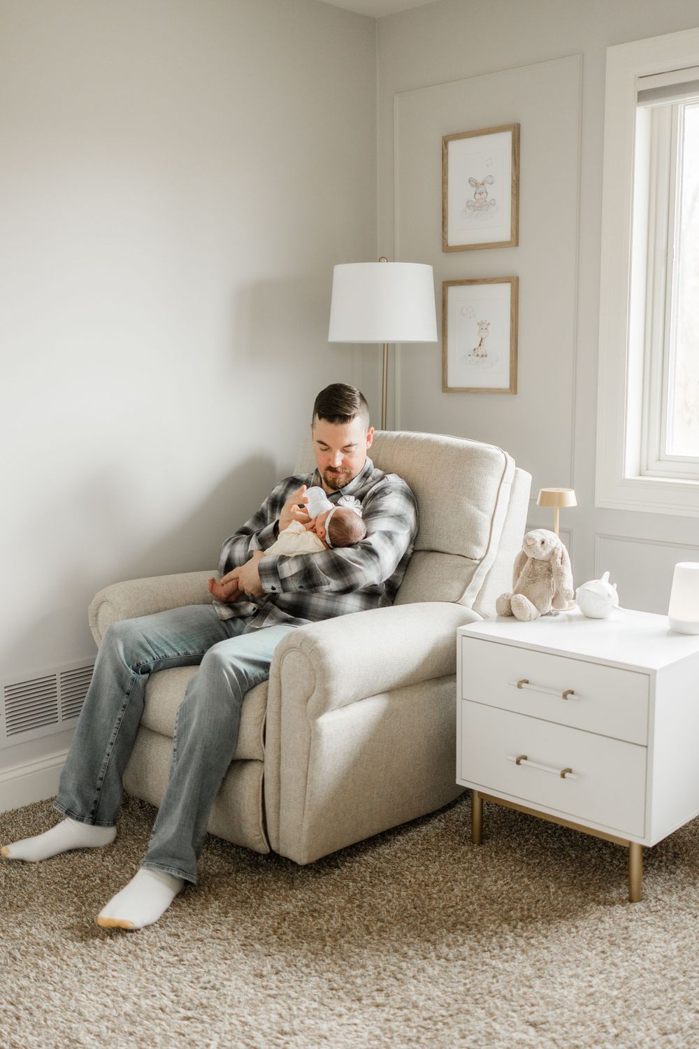 Peaceful nursery scene with caregiver holding baby in plaid shirt while sitting in beige armchair.