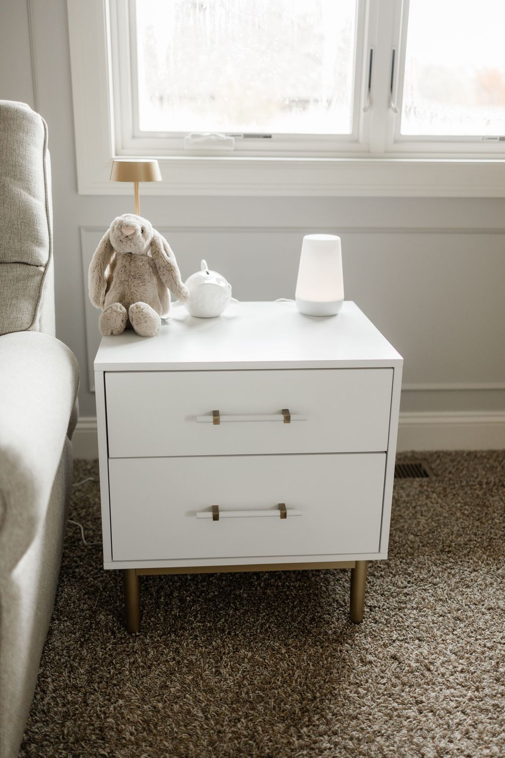 Modern white nursery side table with brass handles and stuffed bunny decoration near a window.