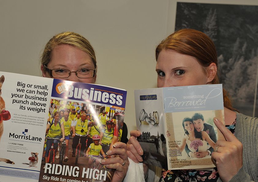 Two women hiding their faces behind business and lifestyle magazines in an office setting.