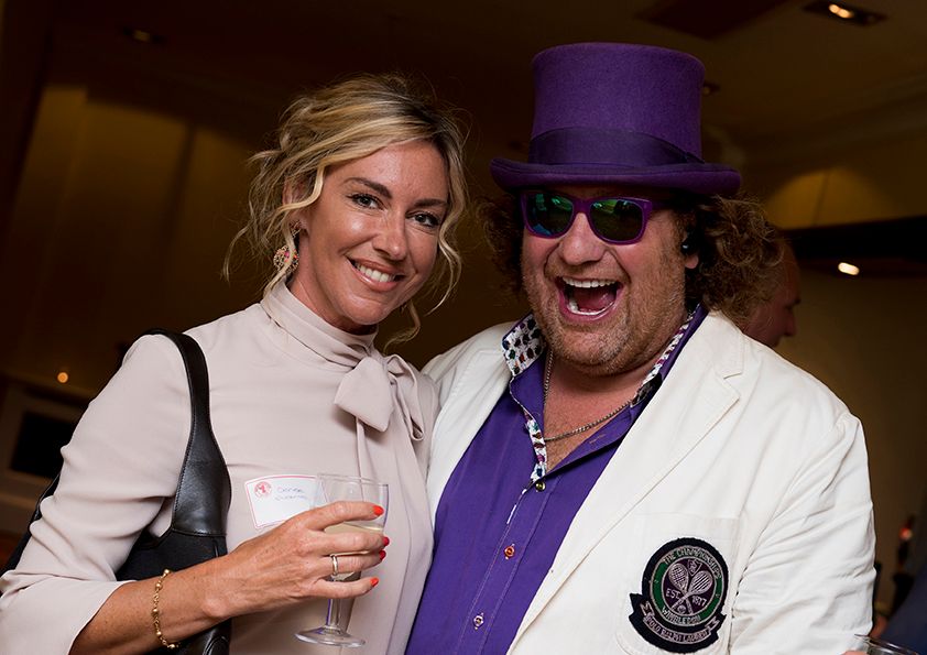 A flamboyant figure in purple top hat and white jacket poses with someone at an indoor event.