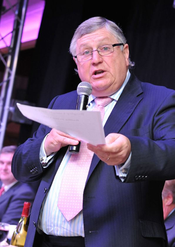 Speaker in blue suit and pink tie addressing audience from stage with microphone.