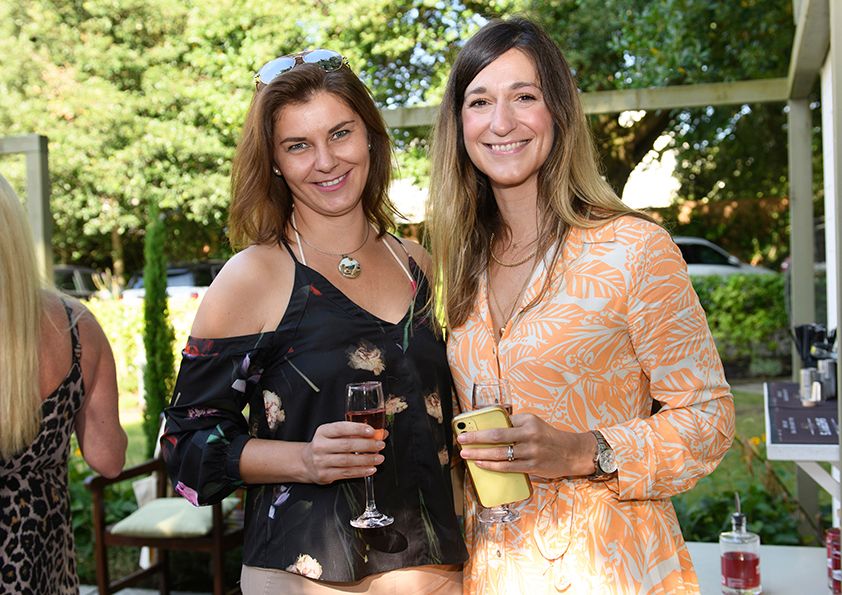 Two friends in summer dresses share drinks at an outdoor garden party.