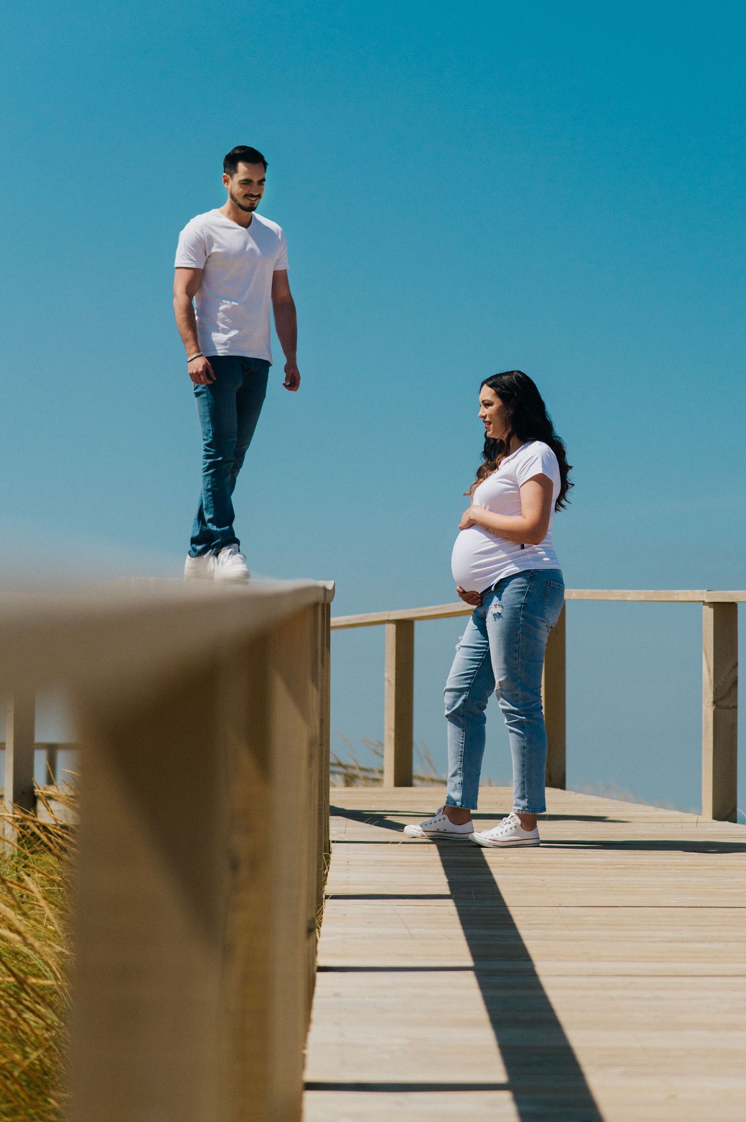 Casual maternity photo of couple standing on wooden boardwalk against blue sky.