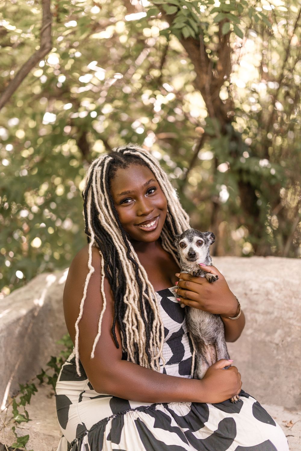 Someone wearing a black and white printed dress poses outdoors while holding a small gray dog in a natural setting.