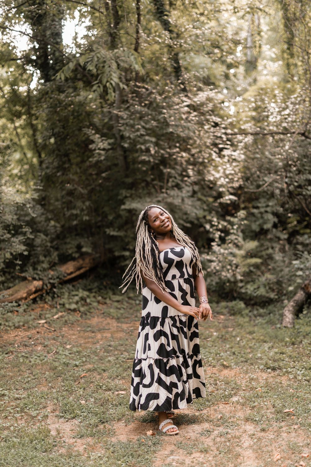 A person wearing a black and white patterned dress sits and poses in a sunny outdoor wooded setting.