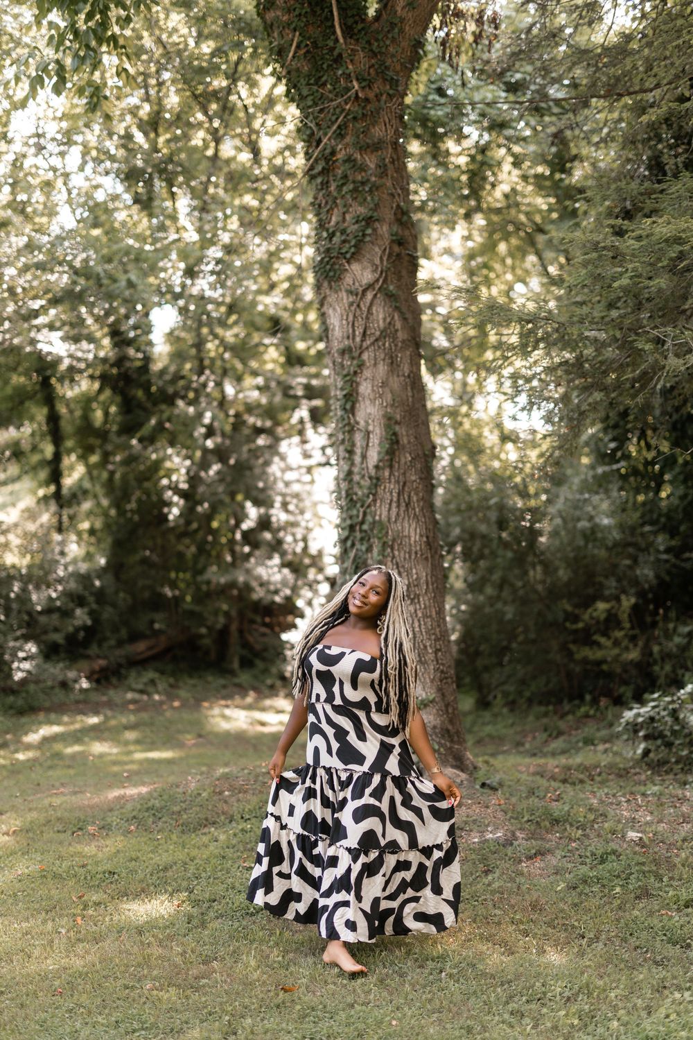 A person in a flowing black and white patterned dress sits serenely against a tree trunk in a peaceful woodland setting.
