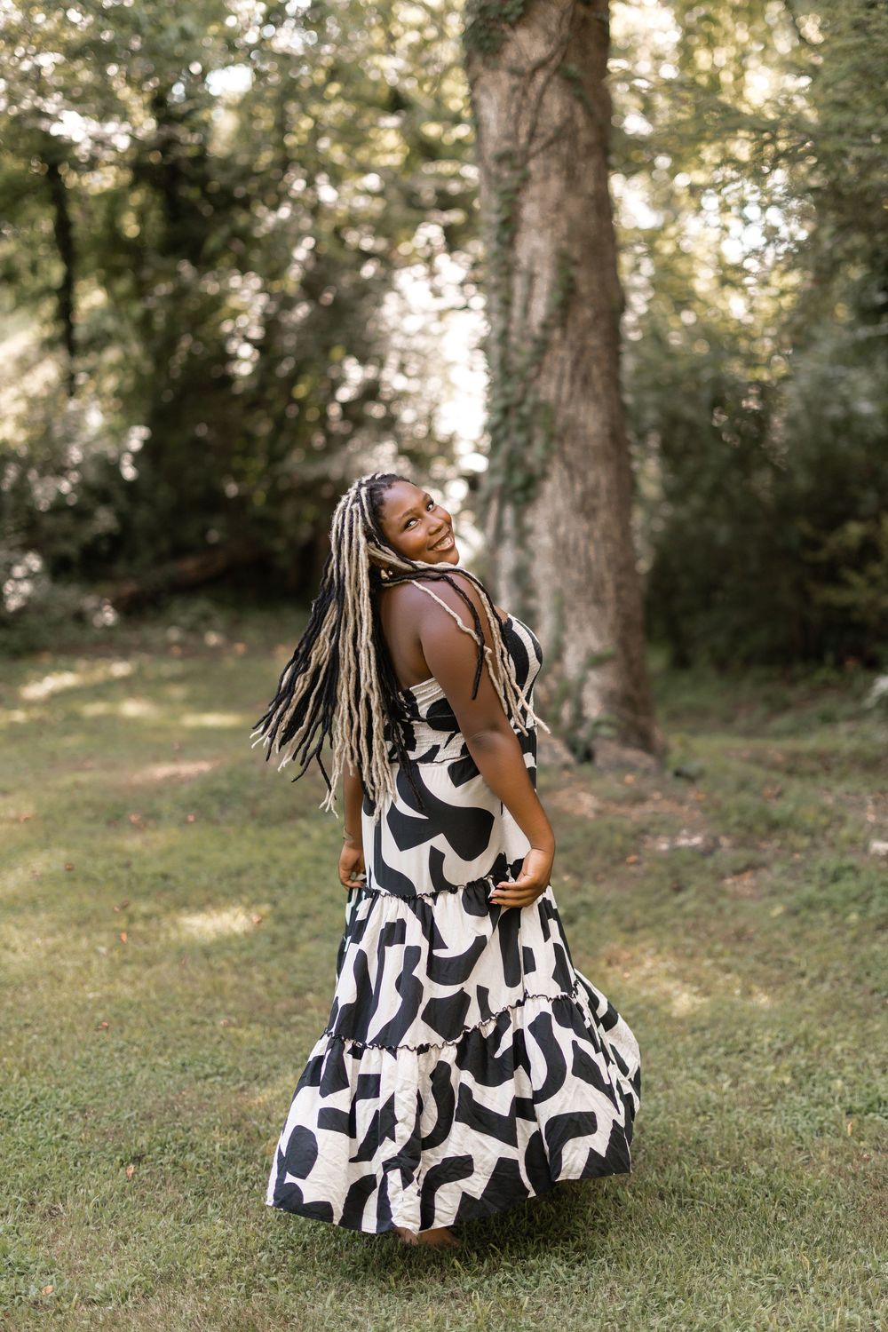 A fashionable figure in a black and white patterned maxi dress twirls joyfully in a sunlit garden setting.
