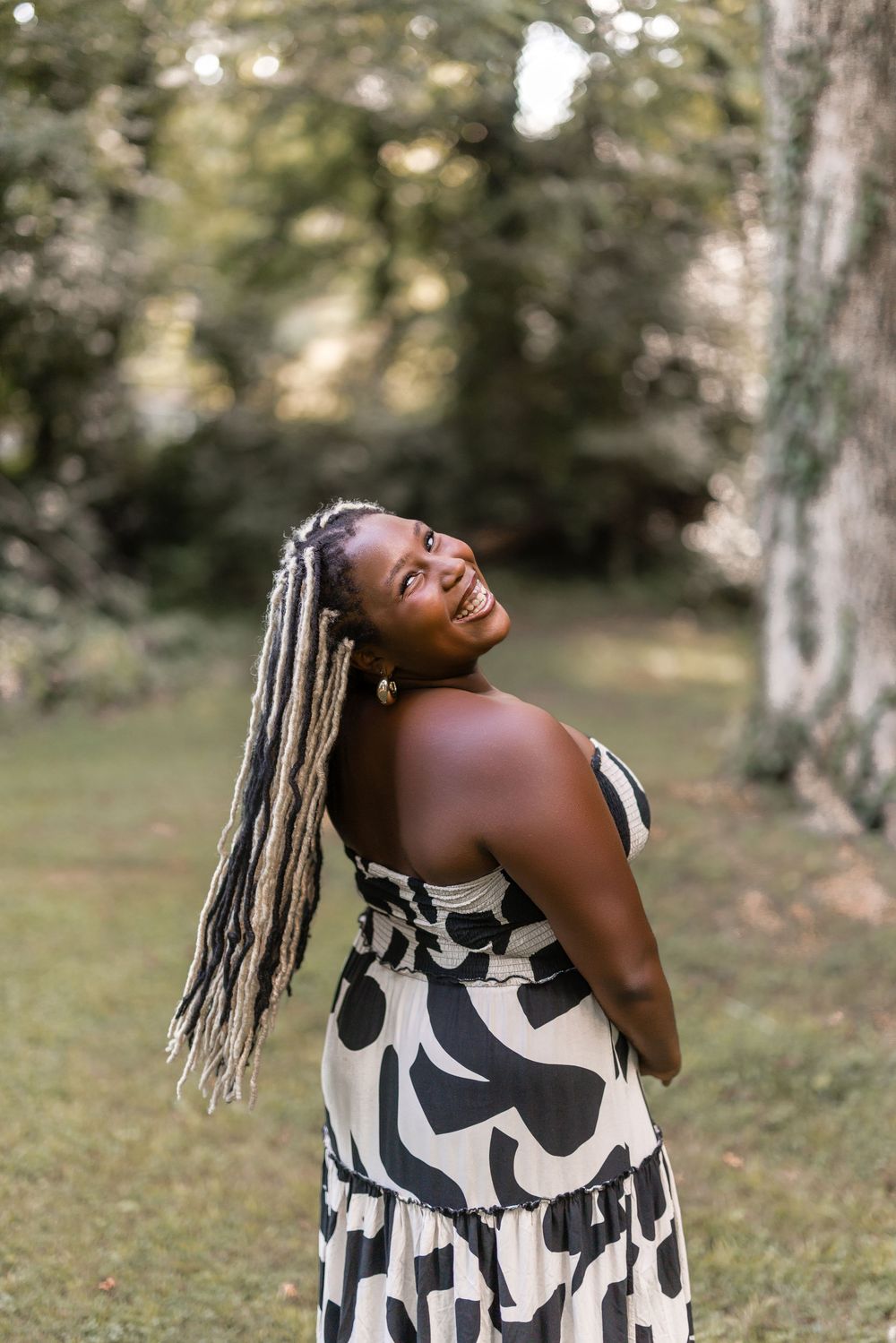 Joyful woman wearing a black and white patterned dress poses outdoors with long braided hair in a garden setting.