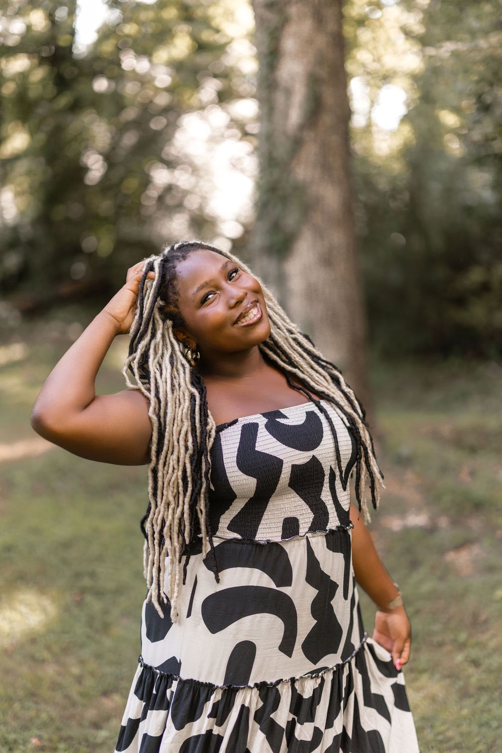 A joyful woman in a black and white patterned dress poses outdoors in a sunlit wooded area.