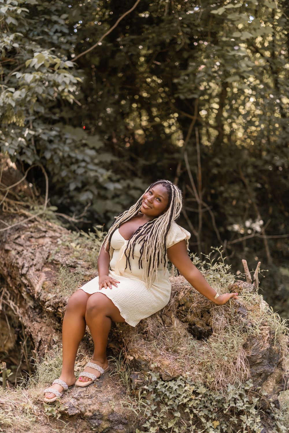 A person wearing a white dress sits gracefully on a rocky slope in a wooded outdoor setting.