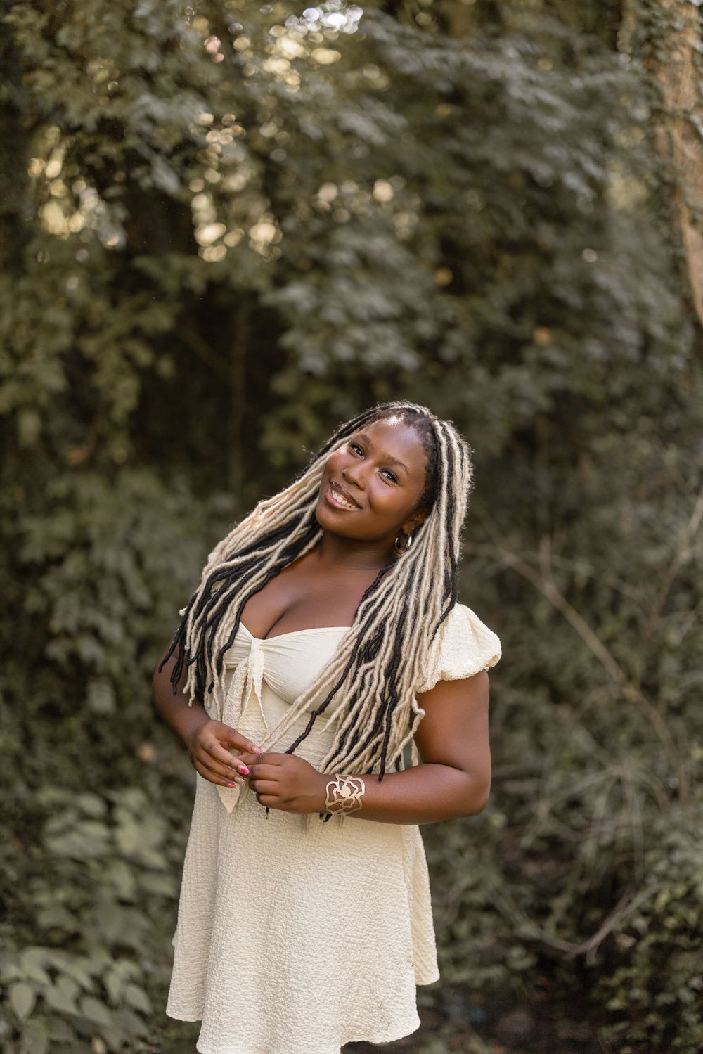 Someone wearing a cream-colored dress poses outdoors with blonde braids while smiling against a natural background.