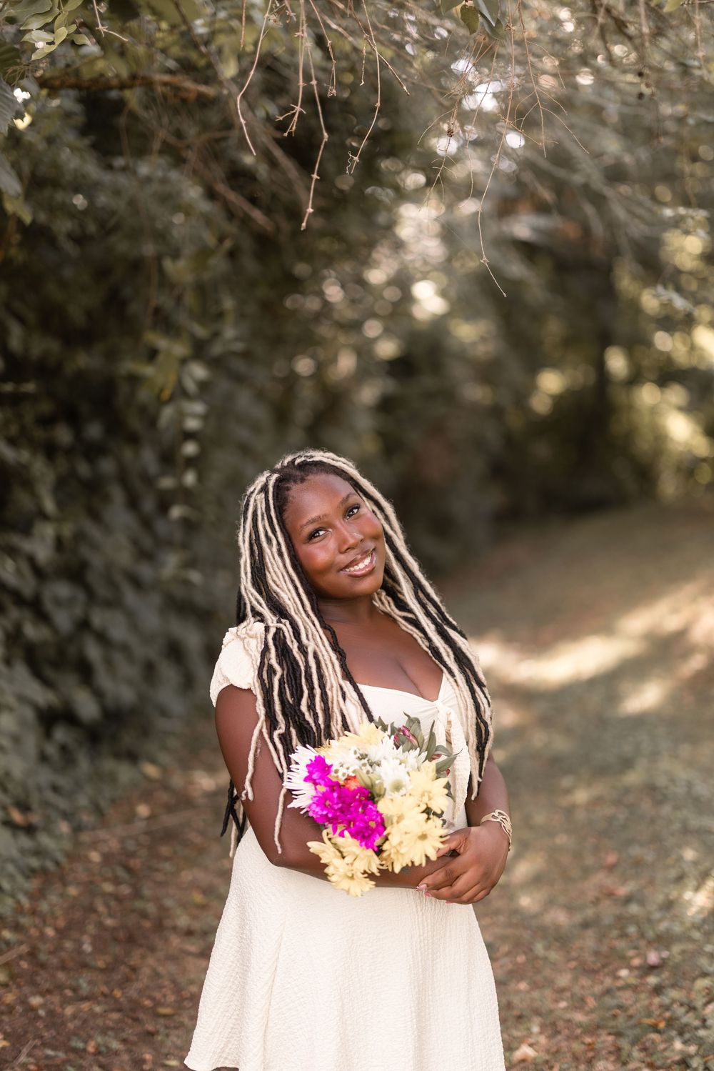 A person in a white dress holds bright pink and yellow flowers while standing in a wooded outdoor setting during golden hour.