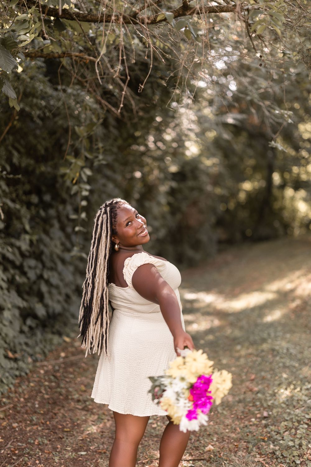 Candid outdoor portrait of woman in white dress holding colorful flowers on a wooded path with dappled sunlight.