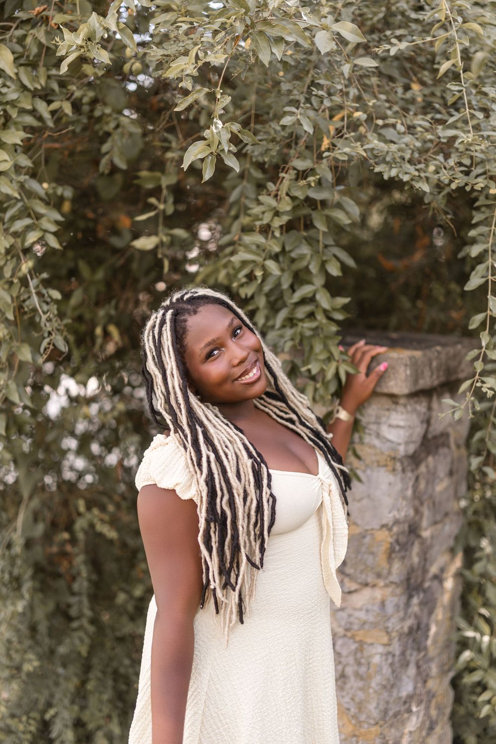 Person in a light dress stands among leafy trees while smiling and posing playfully outdoors.