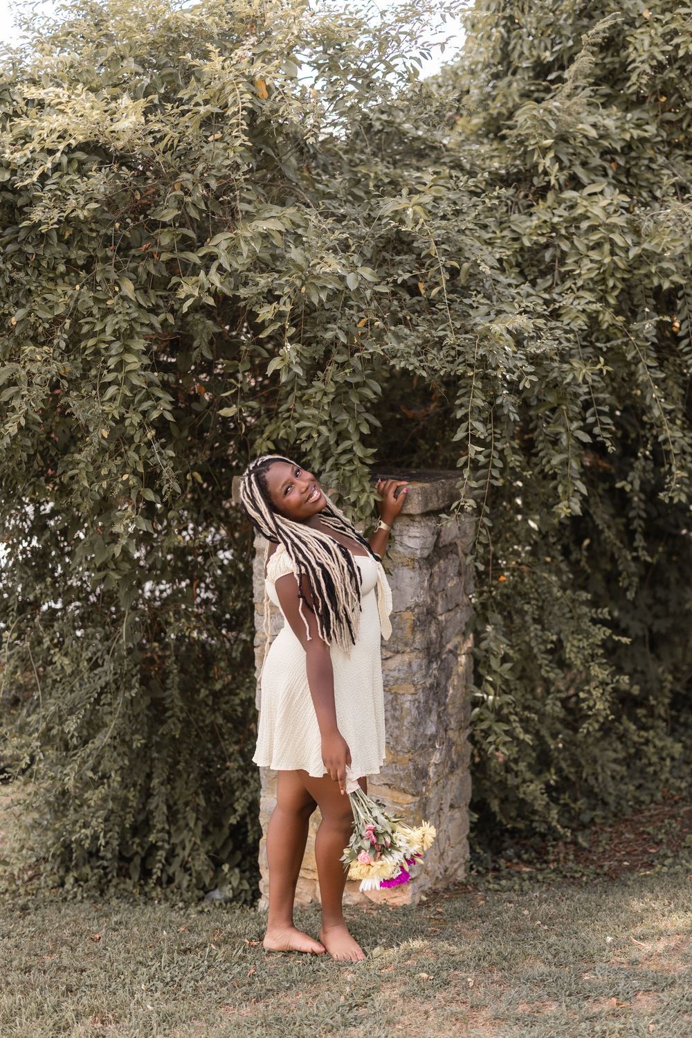A woman in a white dress stands barefoot against a backdrop of lush greenery and trees.
