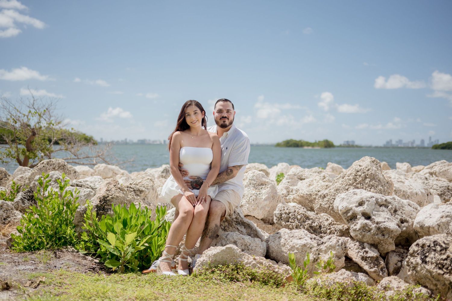 A couple sits together on a rocky beach overlook with the ocean and blue sky in the background.