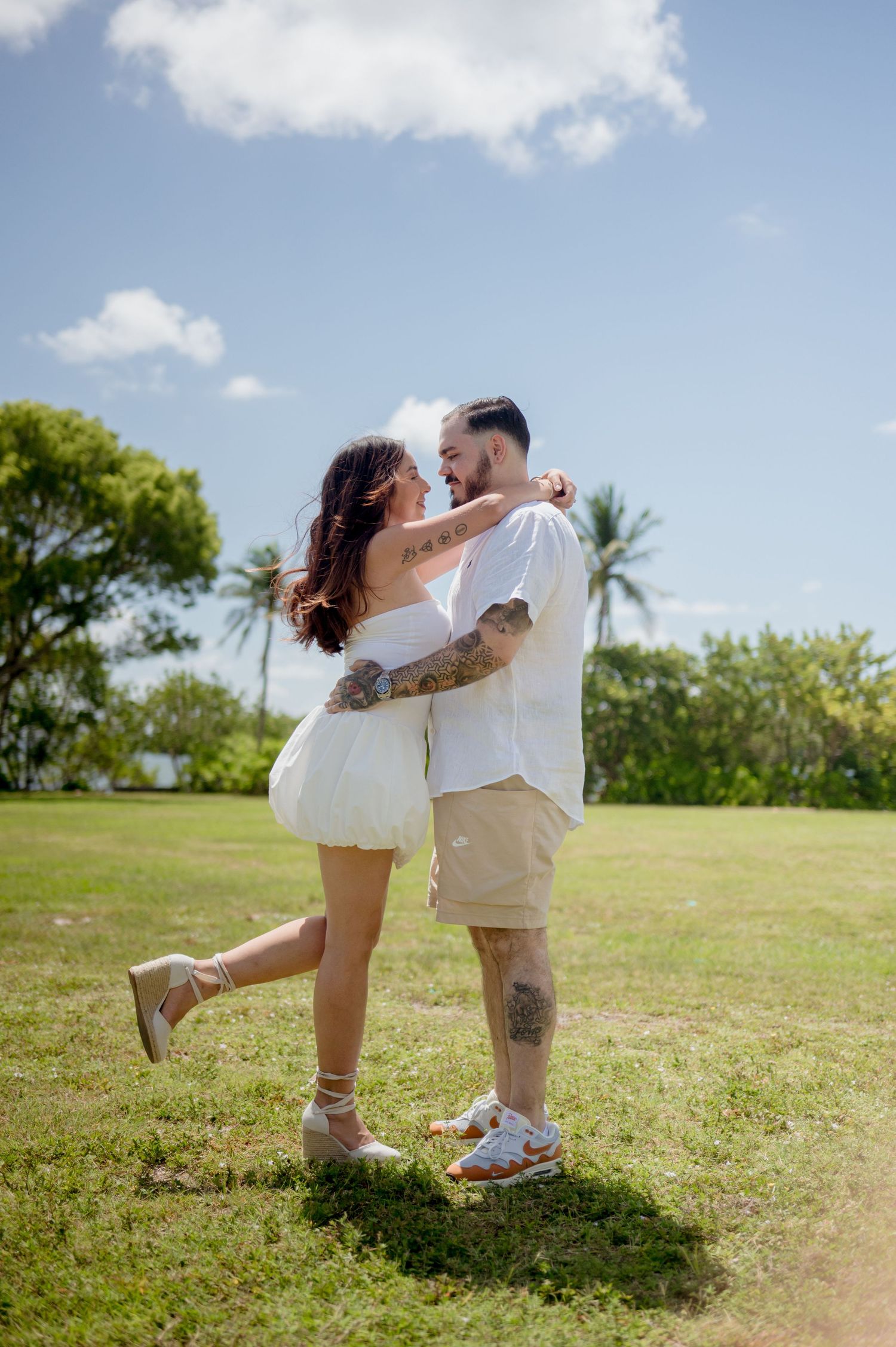 A romantic couple embraces on a sunny day in a grassy field with palm trees and blue sky in the background.