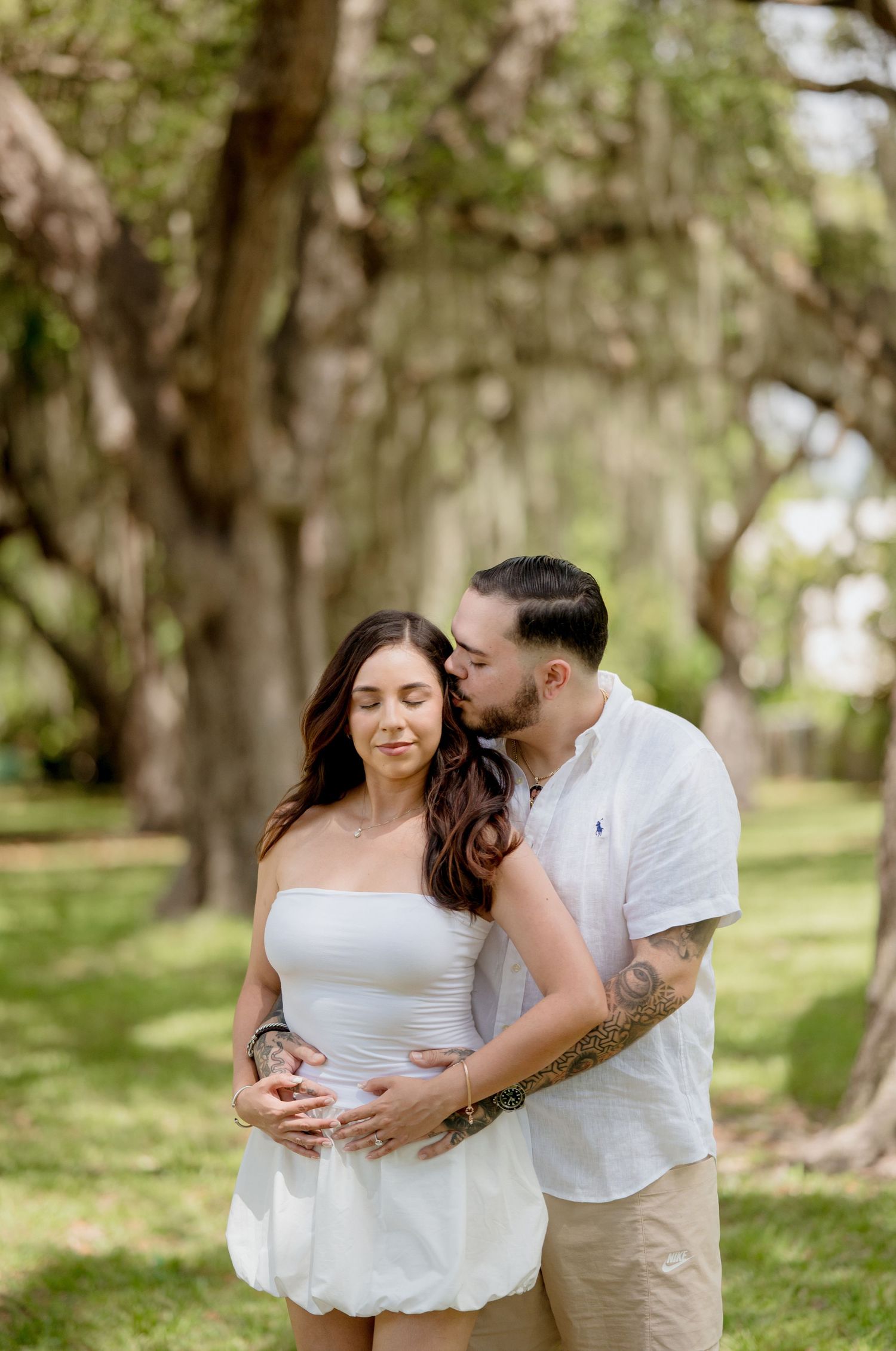 Romantic couple embraces in a sun-dappled park with Spanish moss draped trees creating a dreamy backdrop.