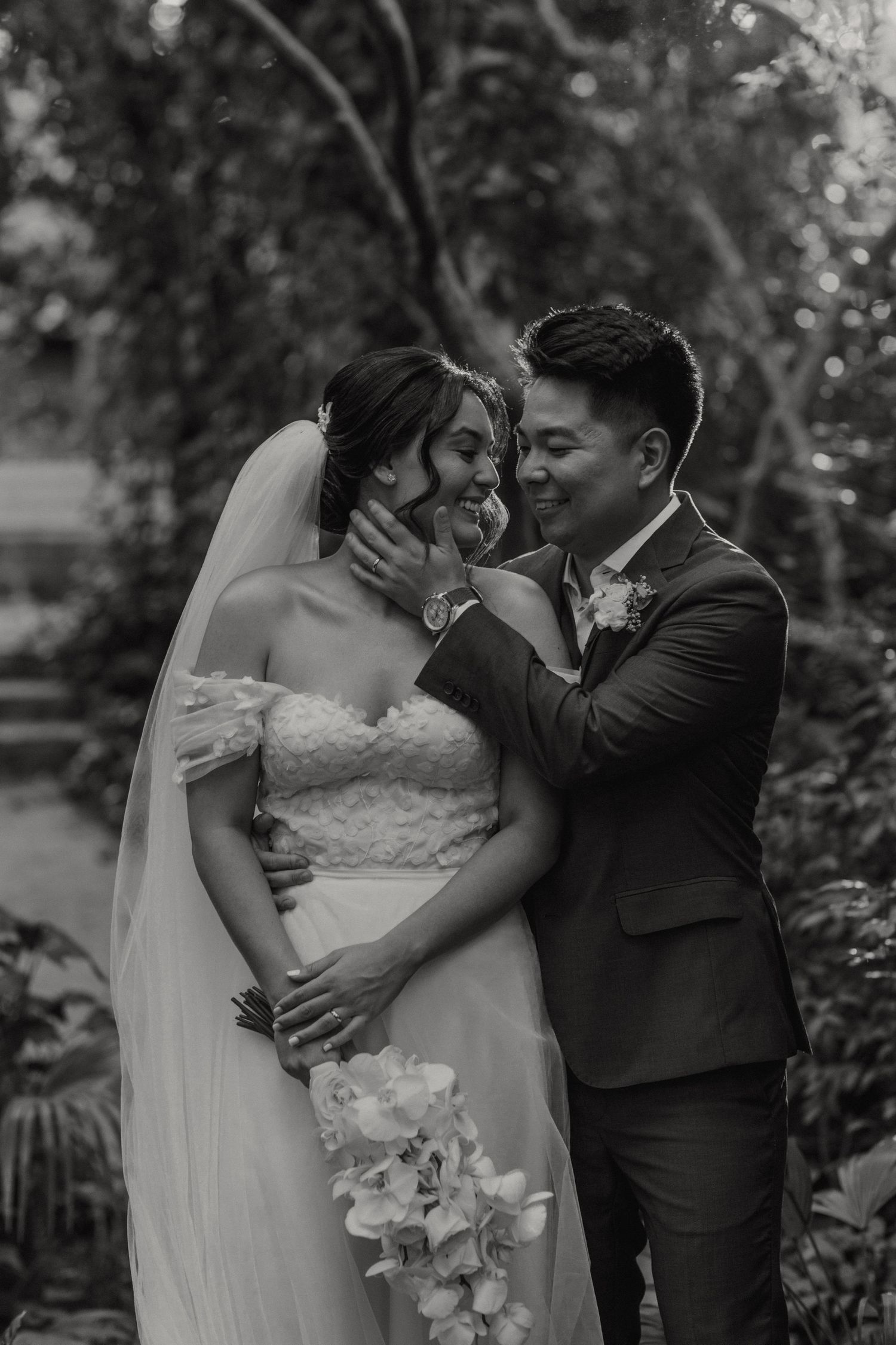 A tender moment between newlyweds sharing a laugh in a garden setting, captured in black and white.
