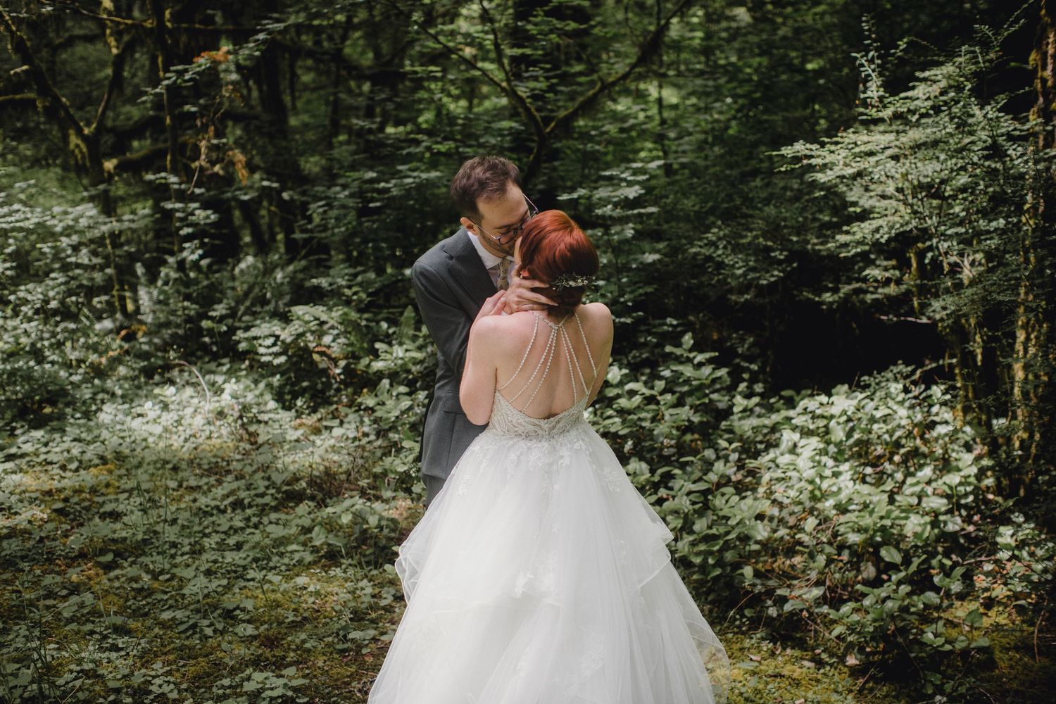 A romantic wedding couple sharing a kiss surrounded by lush green forest and blooming white flowers in the background.