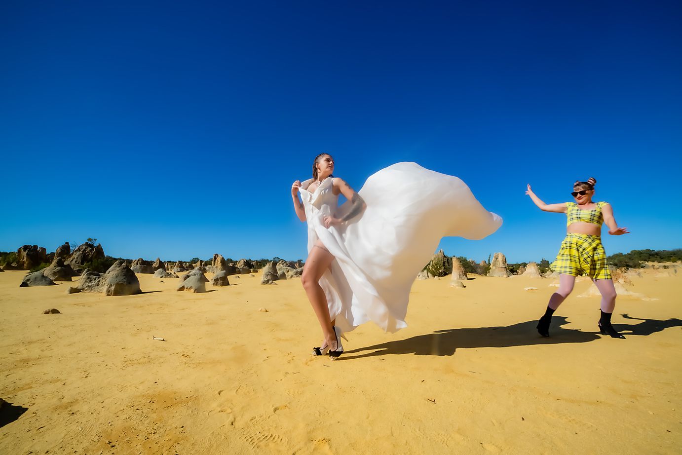 Two people pose playfully among the limestone pinnacles at The Pinnacles Desert in Western Australia on a bright sunny day.