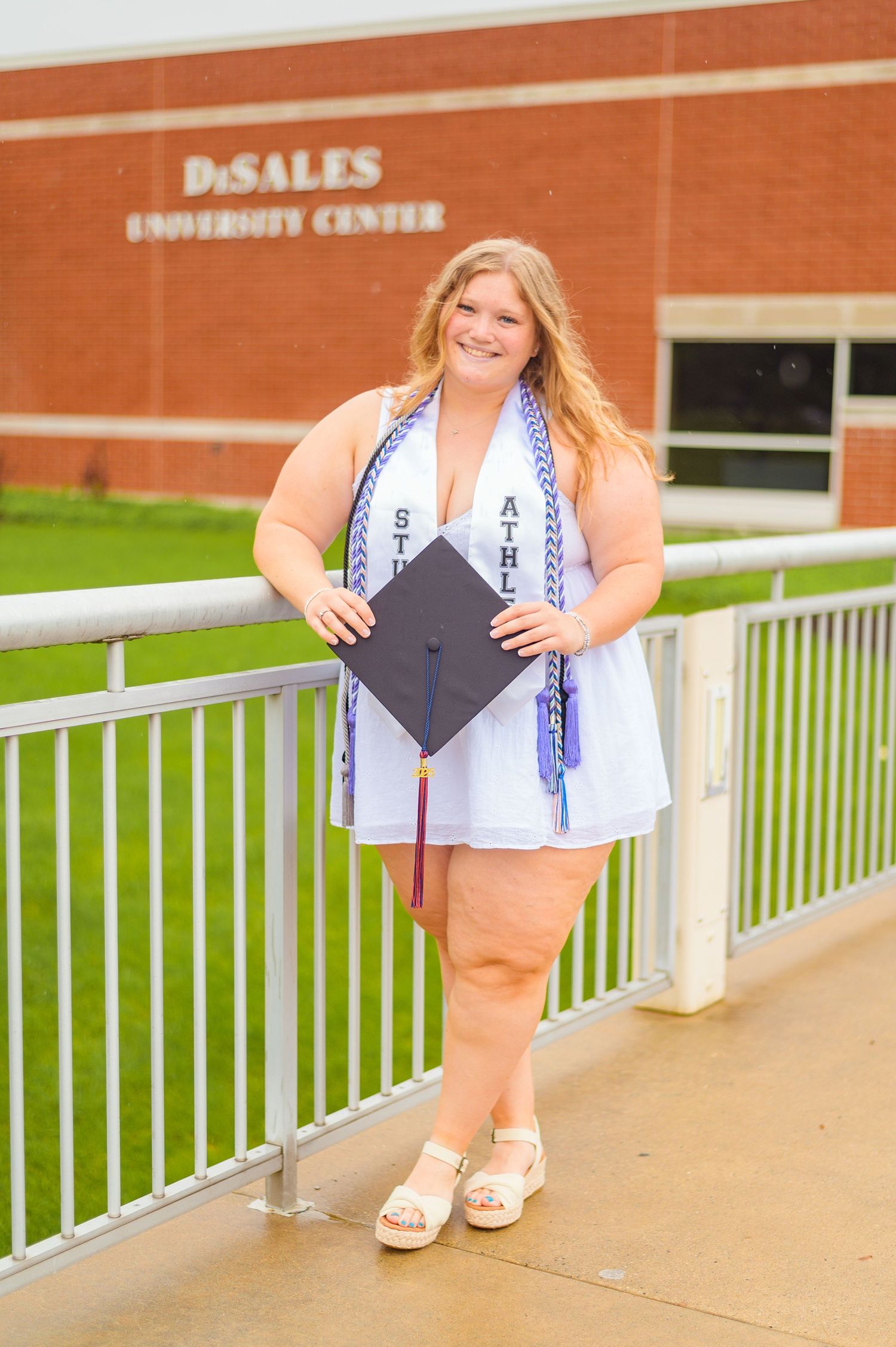 Graduate in white dress poses with diploma outside DeSales University Center building.