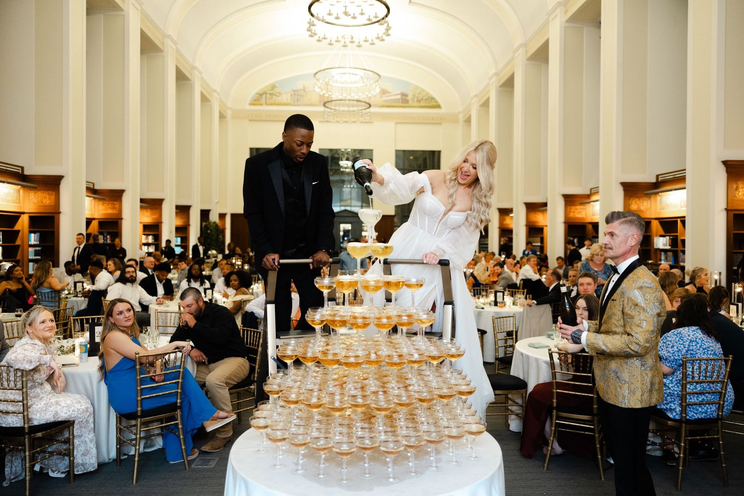 People climbing and pouring champagne on a towering pyramid of glasses at an elegant wedding reception.