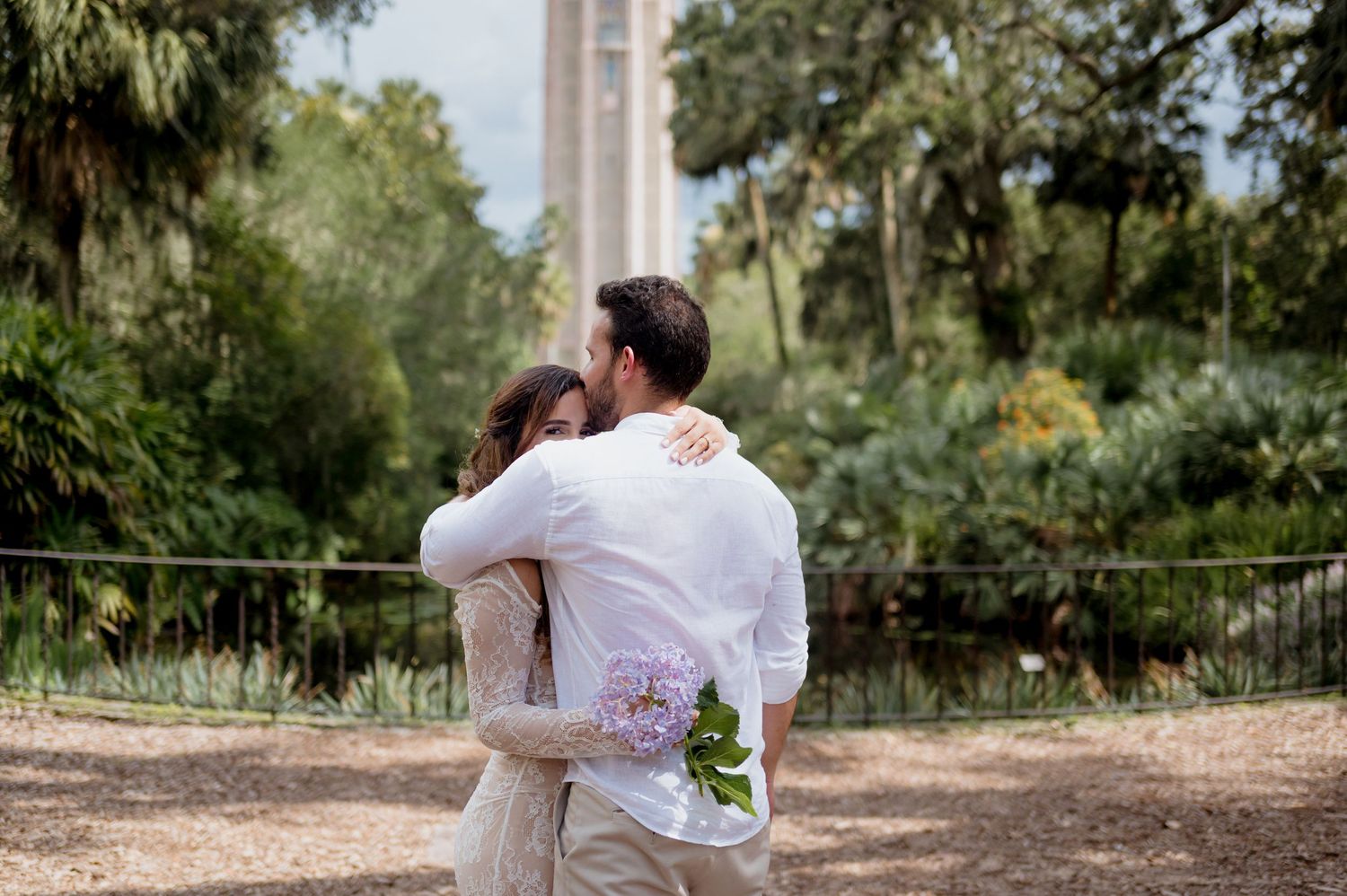 A couple in white clothing share an intimate embrace on a nature path surrounded by lush greenery and palm trees.