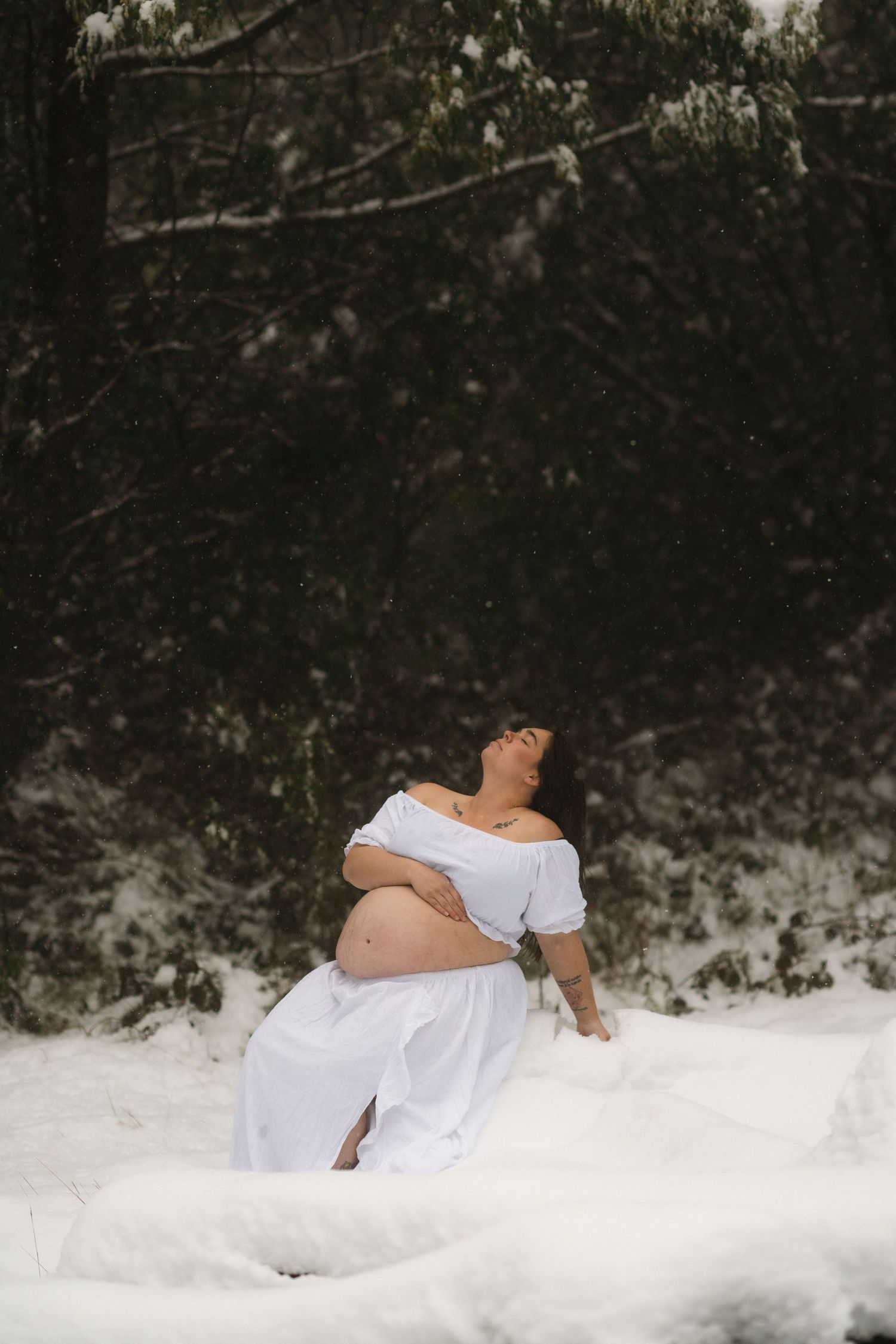 A pregnant woman wearing white clothing lies together in a snow-covered outdoor setting amid dark trees and shadows in Marysville, Victoria