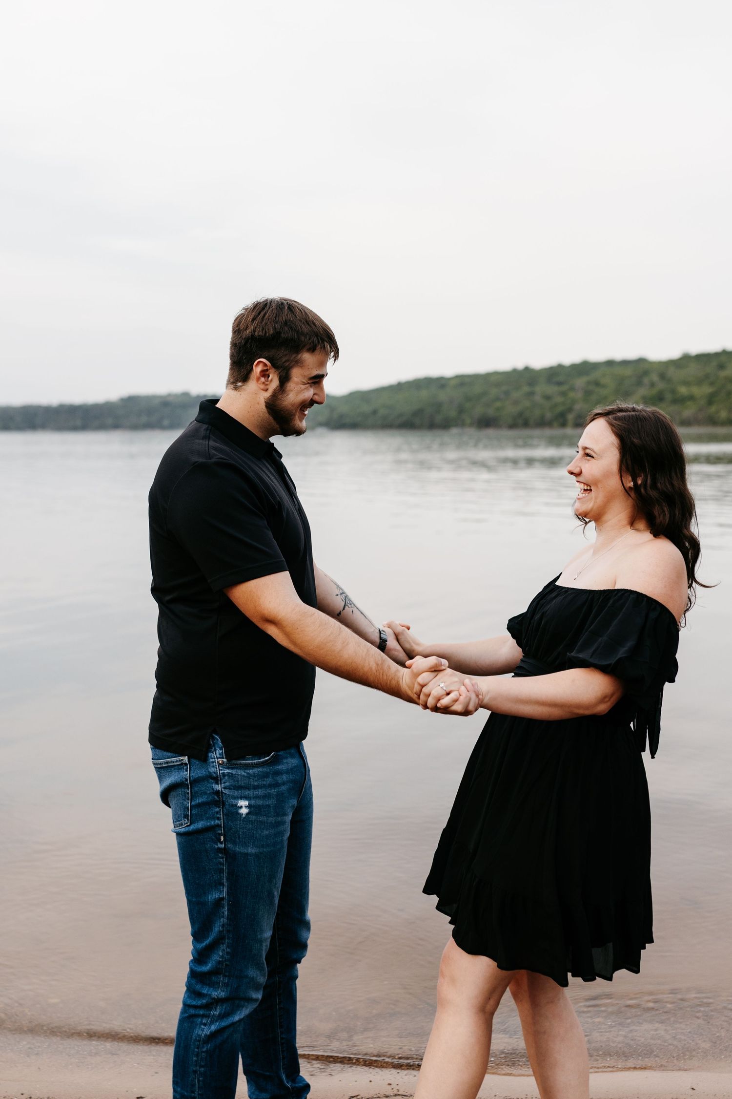 Happy couple in black outfits holding hands and laughing together on a beach with hills and lake in background.