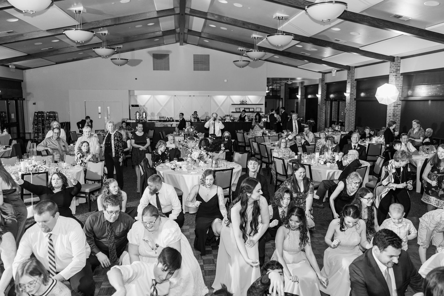 A black and white photograph showing guests dancing and celebrating at an indoor wedding reception venue.