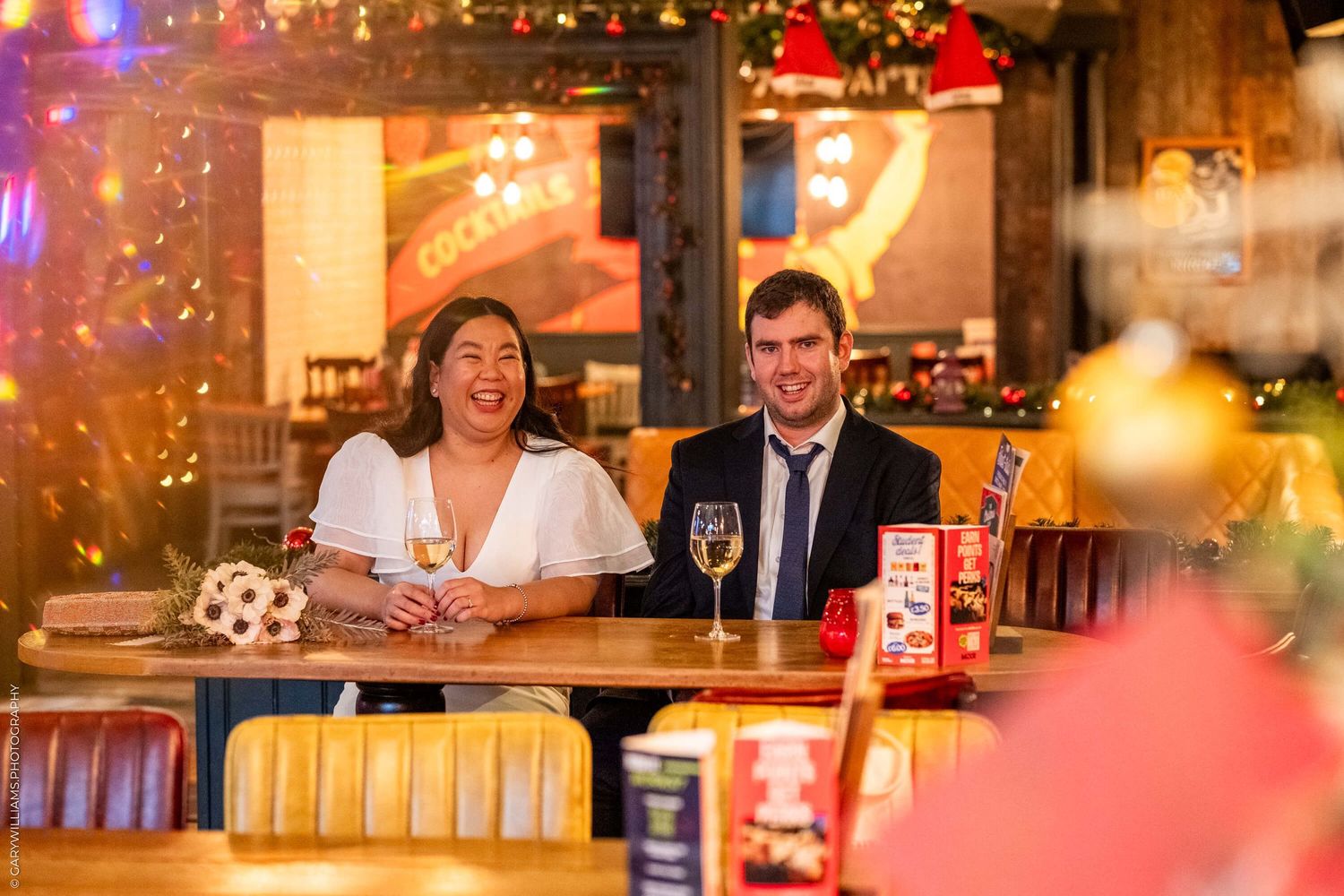 A couple share a joyful moment while sitting at a restaurant table decorated with festive holiday lights and decorations.