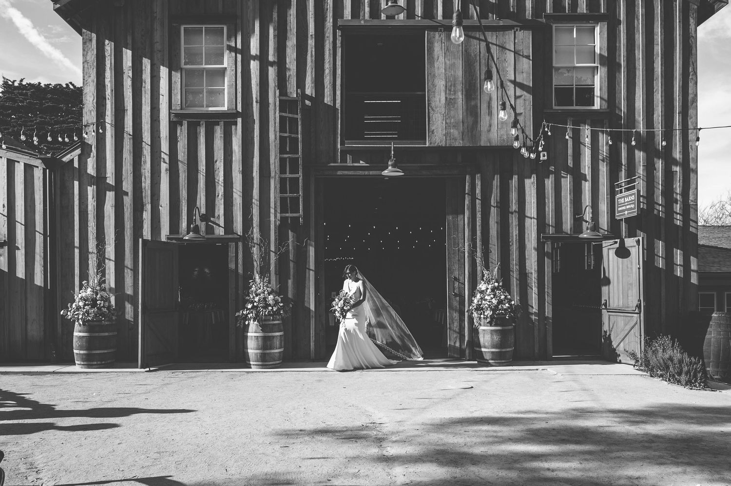 A person in a white dress stands in front of a black and white striped building with potted plants by the entrance.
