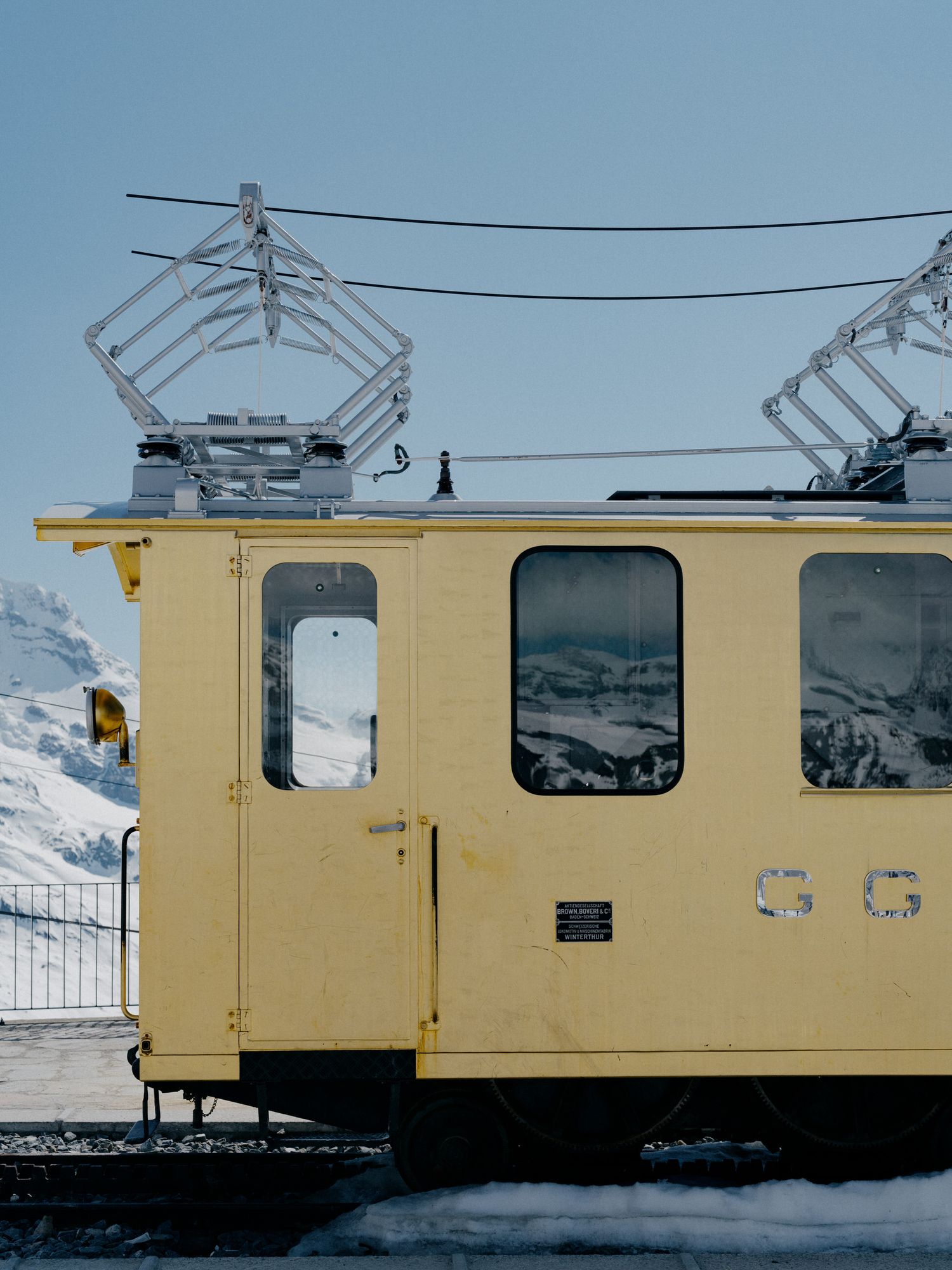 A yellow train car with electric pantographs on top sits in snowy mountain terrain under a bright blue sky.