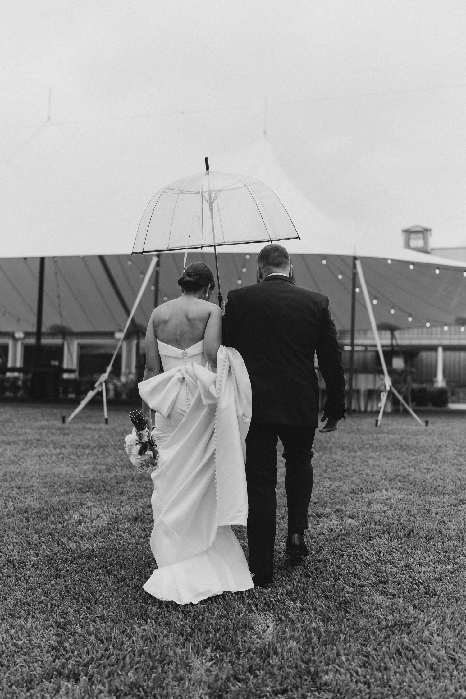 Black and white photo of newlyweds walking under a clear umbrella across a lawn with buildings in background.
