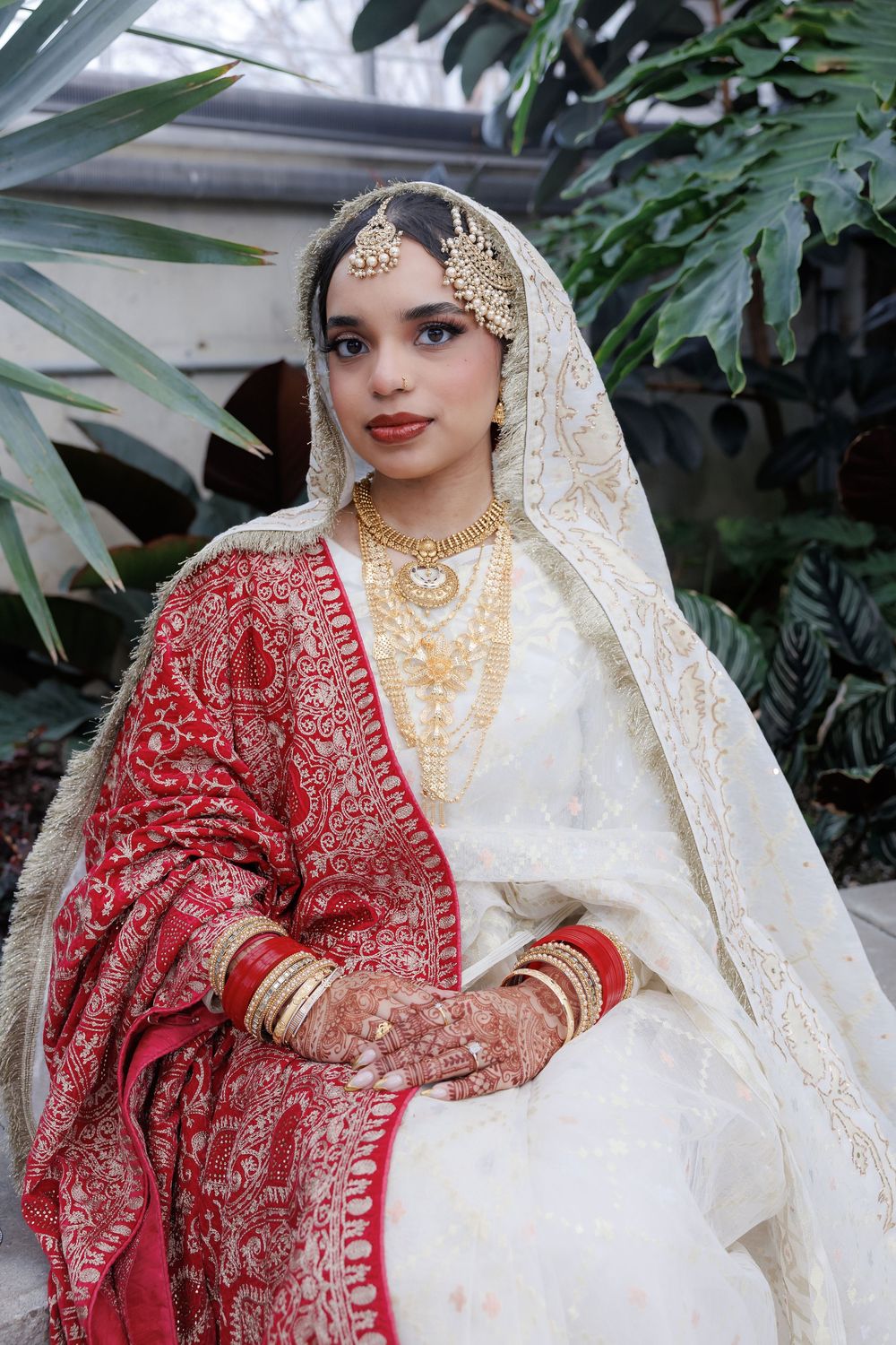 Bride in traditional South Asian wedding attire with red and white saree and gold jewelry against tropical foliage.