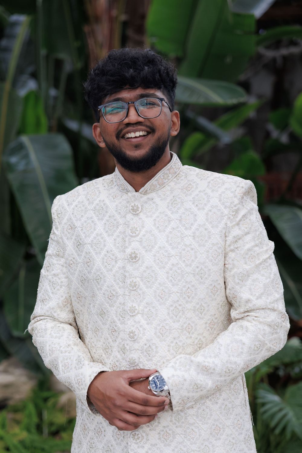 Groom wearing traditional white sherwani smiles against tropical garden background.