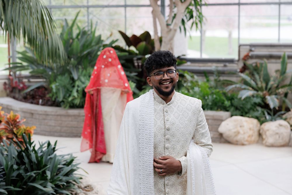 Traditional South Asian groom in white attire standing in a greenhouse with tropical plants.