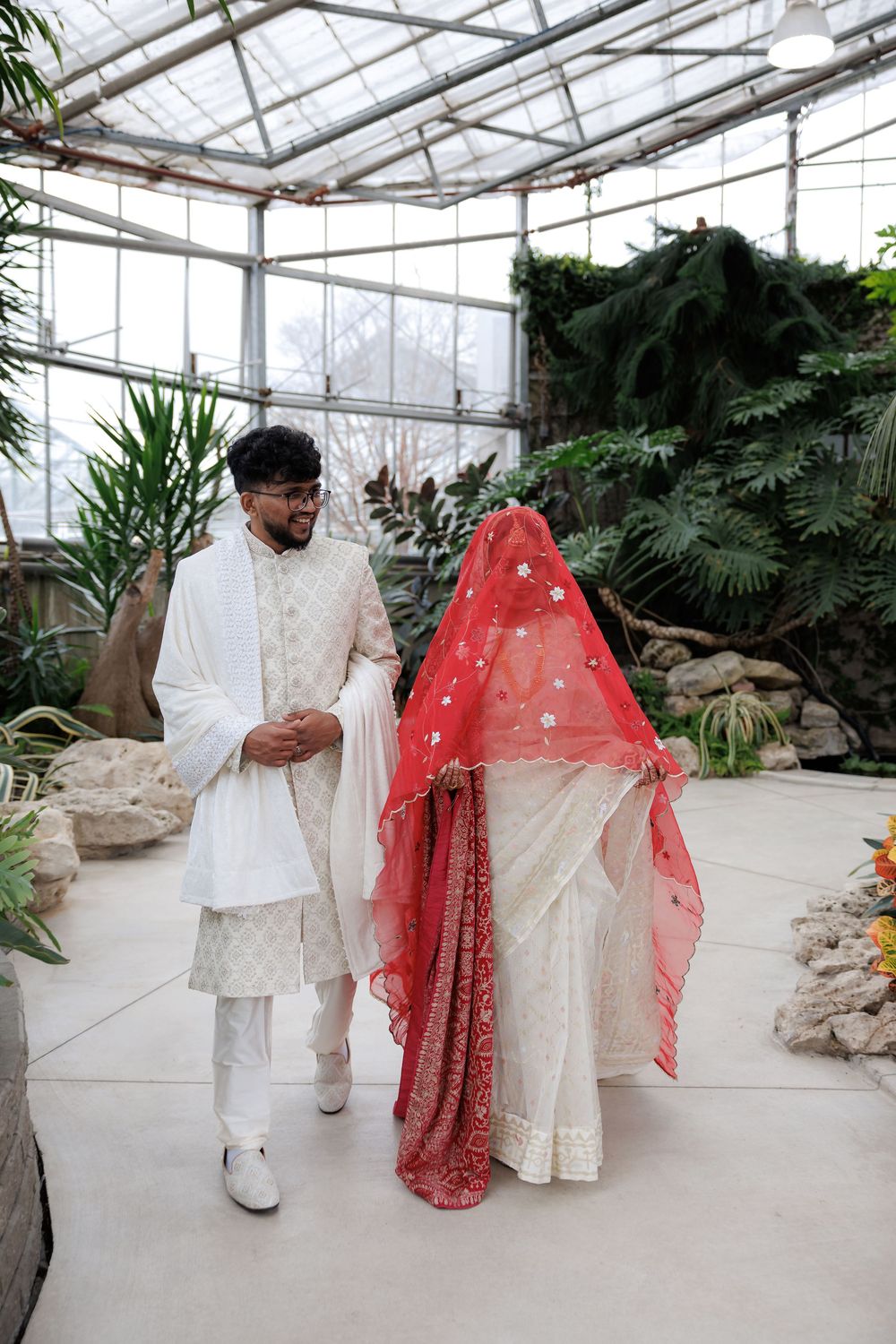Wedding couple in traditional South Asian attire walking through a greenhouse filled with plants.