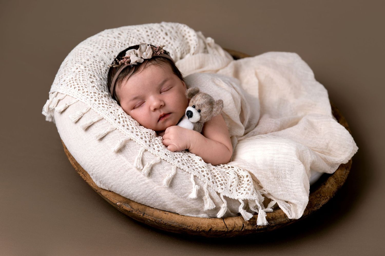 Sleeping newborn baby wrapped in soft white lace blanket resting in round wooden basket on brown background.