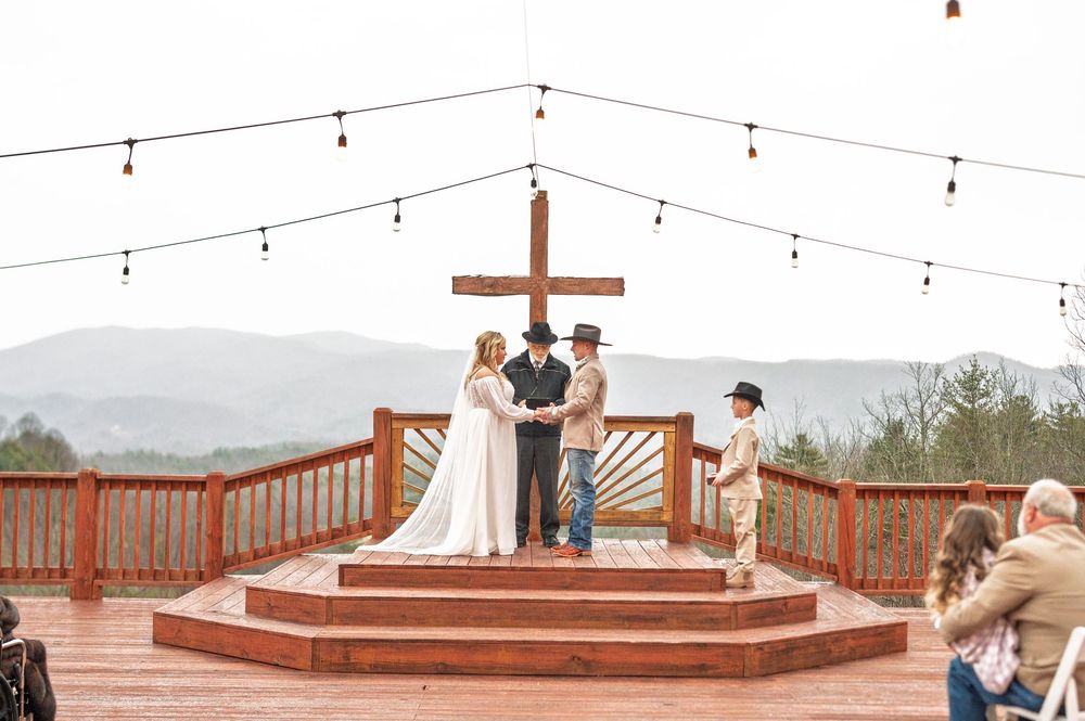 Wedding ceremony setup on a deck with string lights and cross altar against mountain backdrop.